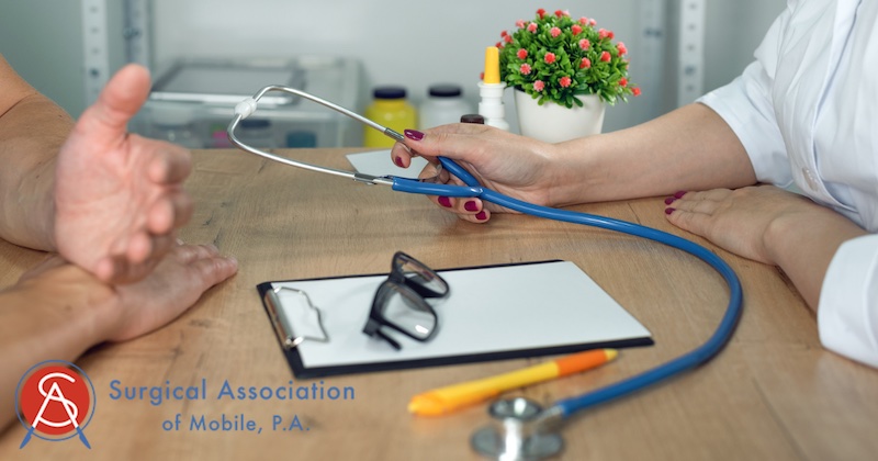 Doctor holding a stethoscope with a clipboard, glasses, pencil at a wood desk with a patient